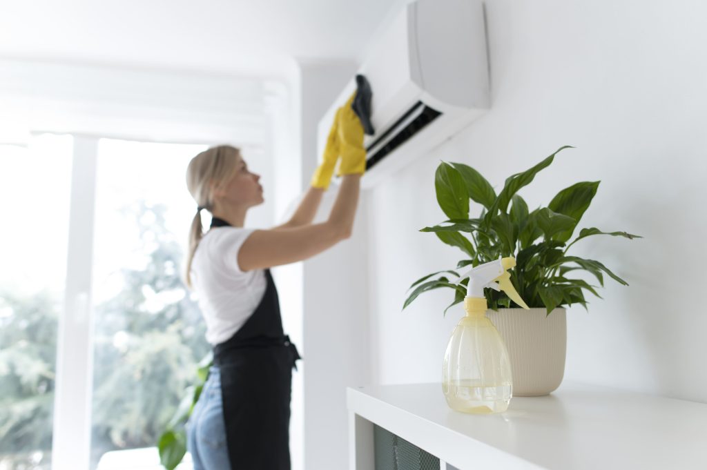 A woman wearing yellow gloves is cleaning an air conditioning unit inside a bright, modern home.