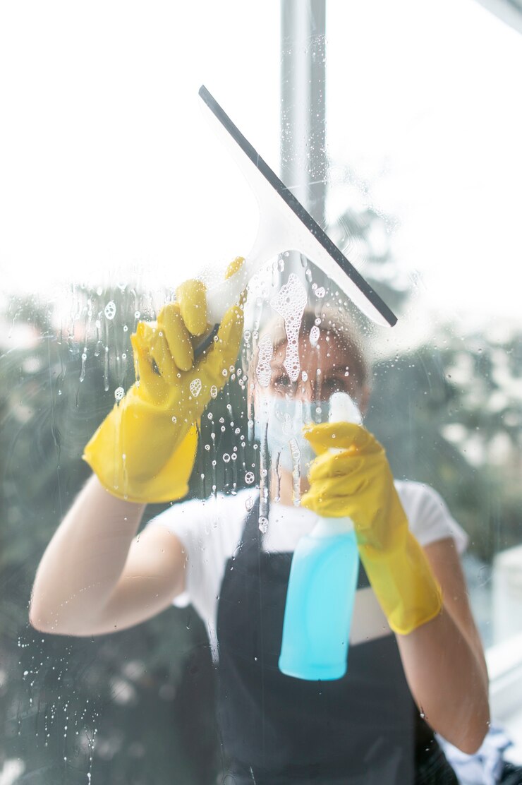 Woman in yellow gloves cleaning a window with a squeegee and spray bottle.