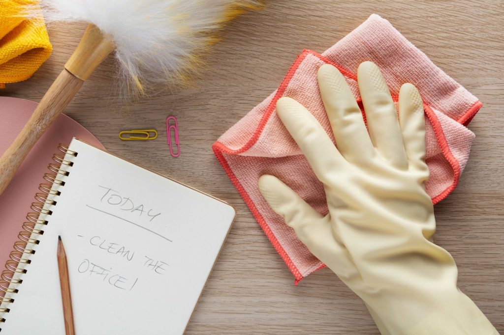 Rubber glove cleaning a desk near a notebook with a cleaning checklist