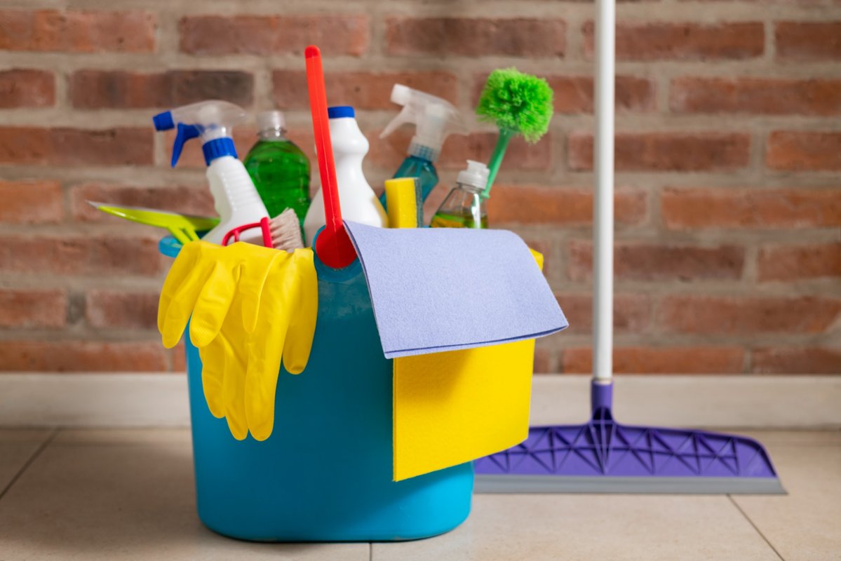 A bucket filled with essential cleaning tools including gloves, sprays, and cloths in front of a brick wall.